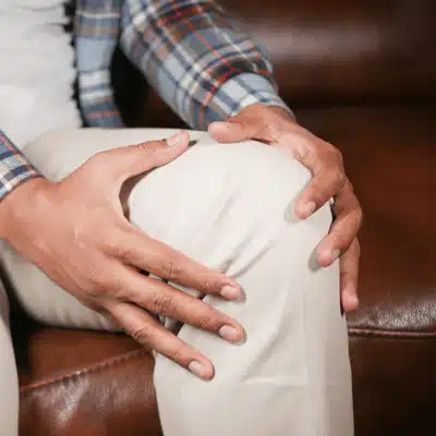 Person holds sore knee while seated on couch awaiting joint specialist evaluation and treatment plan