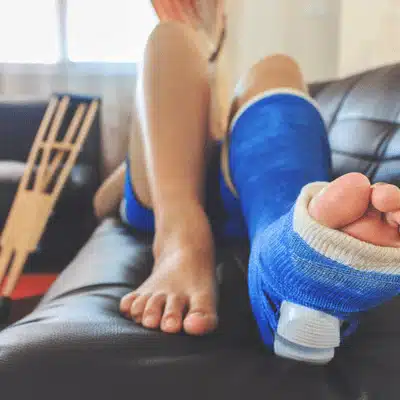 Person rests with blue leg cast on couch, with wooden crutches leaning against nearby wall