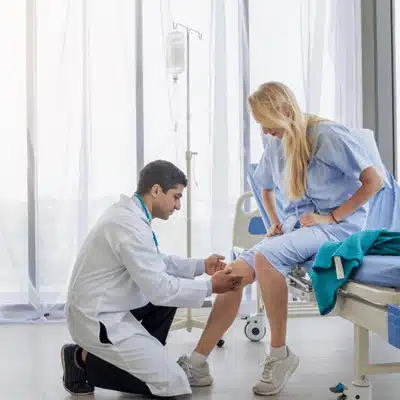 Doctor kneels beside hospital bed, inspecting woman's bandaged knee during recovery assessment after procedure