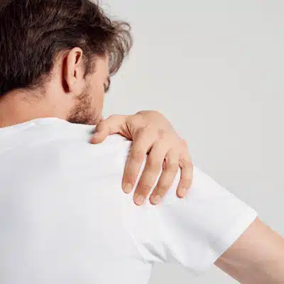 Man grips sore shoulder over white shirt, showing pain signs before shoulder specialist dallas tx visit
