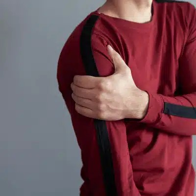 Man holds sore shoulder over red shirt, showing symptoms of rotator cuff tear dallas