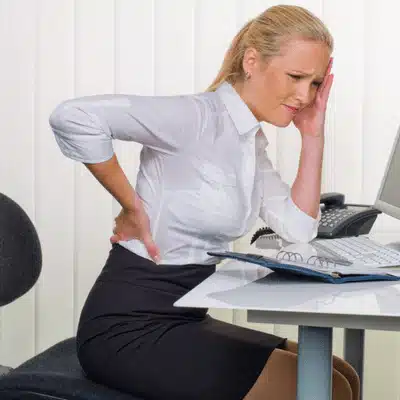 Professional woman seated at desk with back pain possibly needing outpatient hip replacement treatment